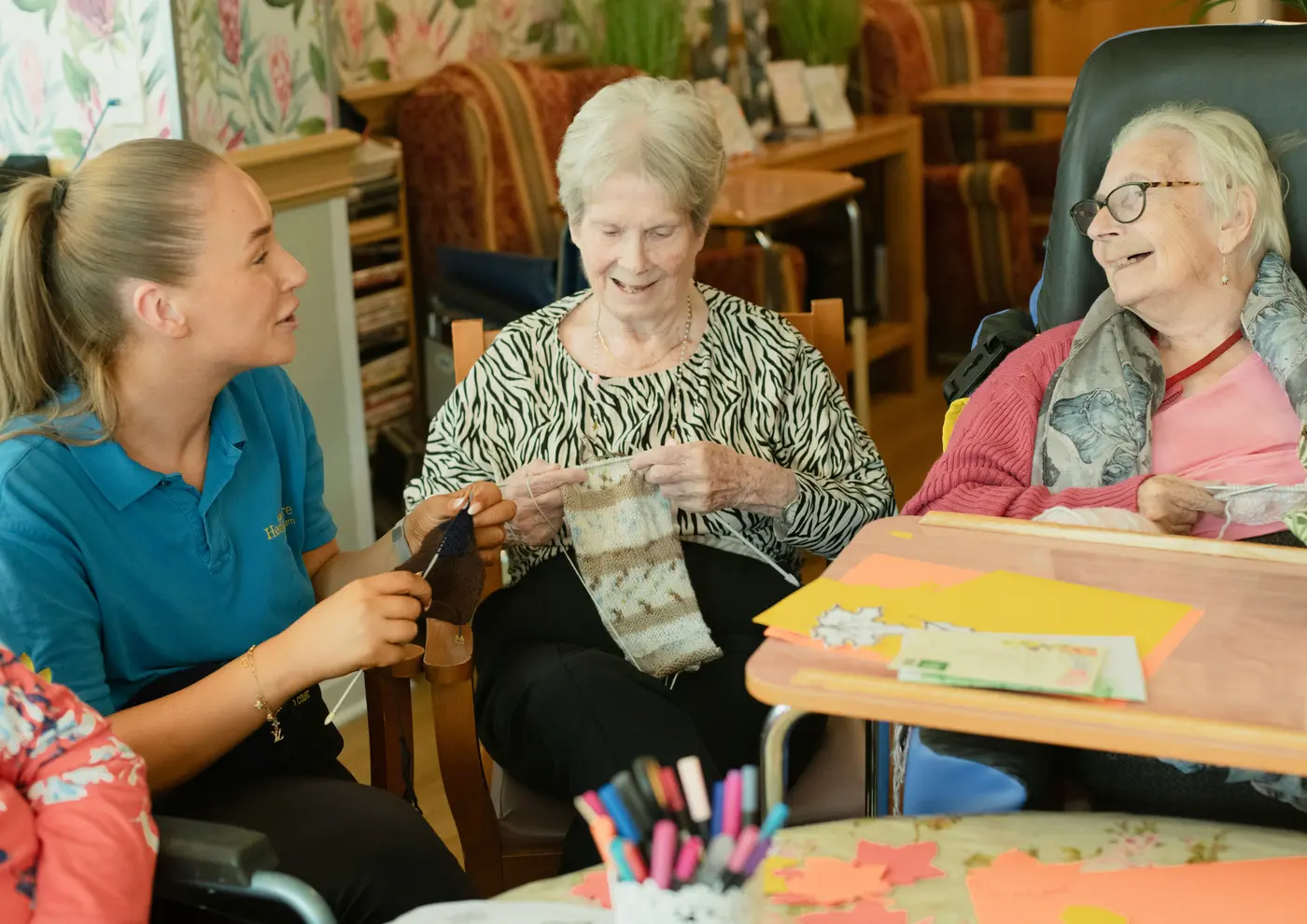 A care worker knitting alongside two older women in a residential lounge, all three smiling.